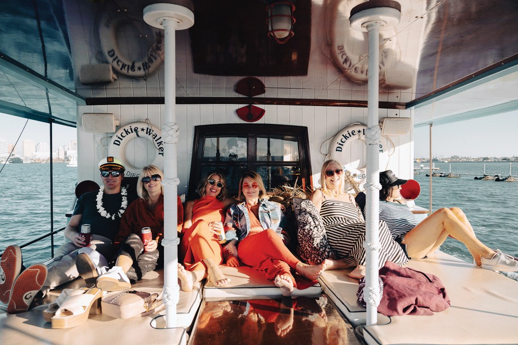 A group of smiling passengers relaxing on the cushioned aft lounge deck of the classic yacht Dickie Walker, framed by white posts, custom life rings with the boat's name, and open water in the background.