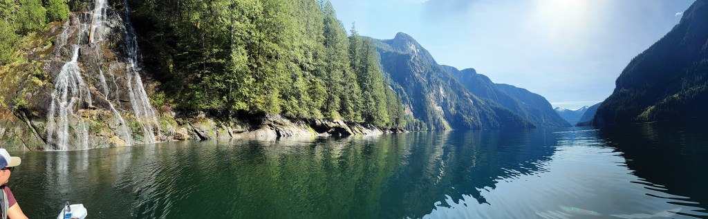 A panoramic view taken from the water in Princess Louisa Inlet, showing a waterfall cascading down rocky cliffs directly into calm fjord waters, bordered by evergreen forest and steep mountain ranges receding into the bright horizon.