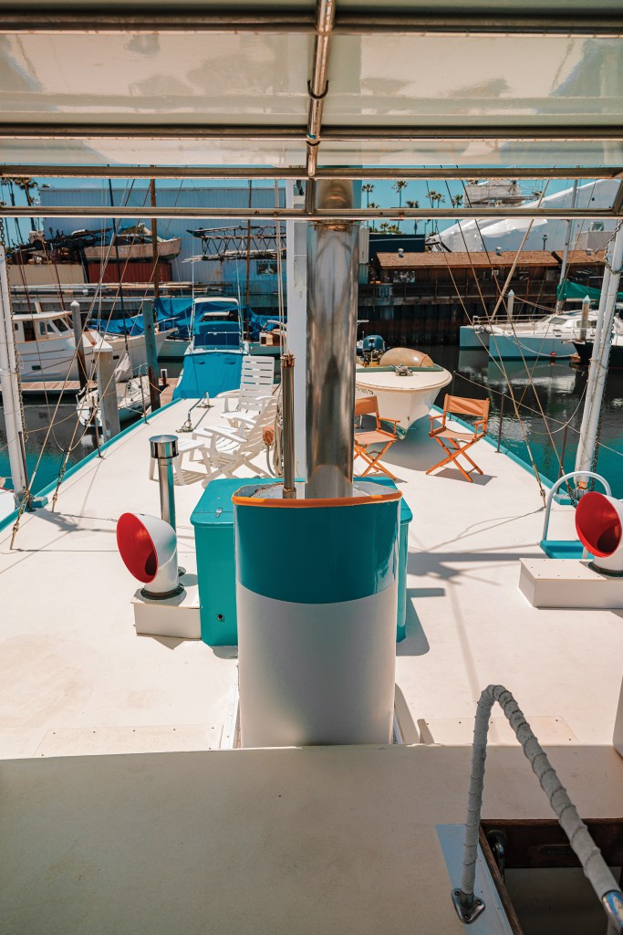 View looking out across the upper sun deck of the classic wooden yacht Dickie Walker, highlighting a blue-and-white exhaust stack, red cowl ventilators, lounge chairs, and a small tender boat docked in the marina background.
