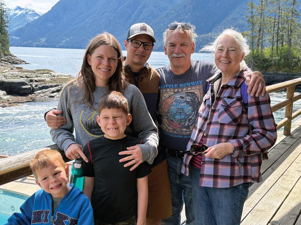 A family portrait of four adults and two young boys smiling together on a wooden deck overlook, with blue waters and steep coastal mountains visible in the background.
