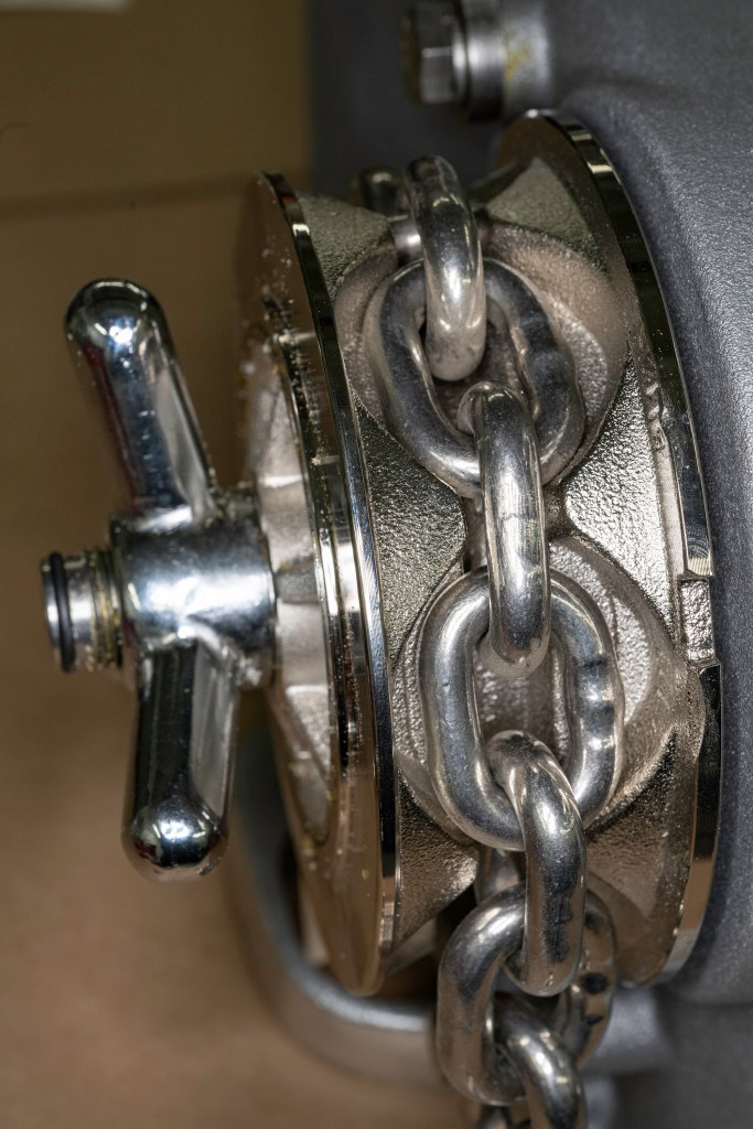 Stainless-steel anchor chain links cleanly aligned inside the pockets of a windlass gypsy, secured by a chrome clutch wing nut.