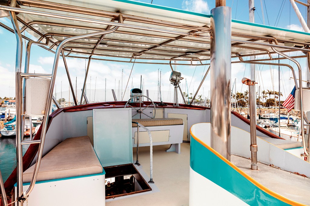 Upper deck helm station of the classic wooden yacht Dickie Walker, featuring a stainless steel bimini frame, secondary steering wheel, cushioned bench seating, access hatch, and a clear view across the marina.