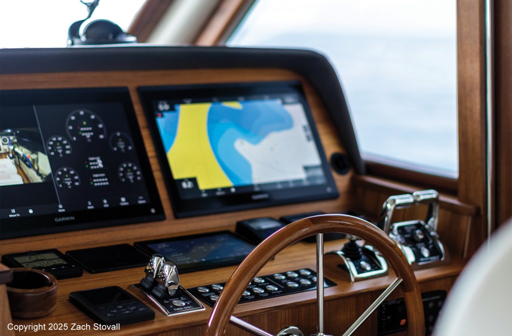 An interior view of a classic teak-accented yacht helm featuring modern Garmin navigation screens, electronic engine controls, a traditional wooden steering wheel, and joystick docking controls.