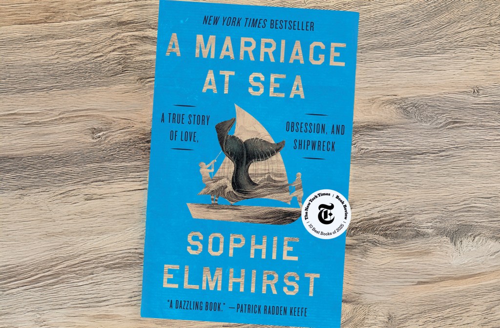 feat_A Marriage At Sea The bright blue front cover of the book A Marriage at Sea by Sophie Elmhirst, featuring an illustration of a whale's tail breaching near a small sailing boat, resting on a light wood surface.