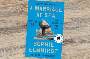 The bright blue front cover of the book A Marriage at Sea by Sophie Elmhirst, featuring an illustration of a whale's tail breaching near a small sailing boat, resting on a light wood surface.