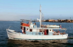 feat_Dickie Walker The restored 65-foot classic wooden yacht Dickie Walker cruising through blue waters near San Diego, featuring a white hull, light blue and wood trim, passengers on deck, and a tall mast under a clear sky.