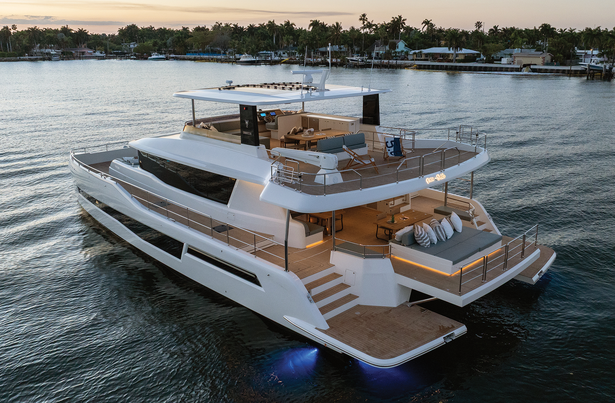 An aerial high-angle view of a white GranOcean W-72 luxury power catamaran anchored in calm coastal waters at dusk, highlighting its expansive multi-deck outdoor living spaces, swim platform, flybridge, and ambient LED deck lighting.