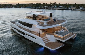 An aerial high-angle view of a white GranOcean W-72 luxury power catamaran anchored in calm coastal waters at dusk, highlighting its expansive multi-deck outdoor living spaces, swim platform, flybridge, and ambient LED deck lighting.