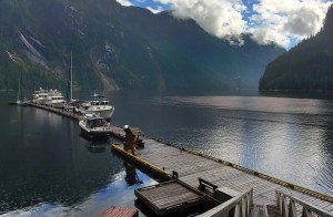 A scenic overhead view looking down a long, wooden floating dock at Princess Louisa Marine Provincial Park in British Columbia, with several powerboats and sailboats moored along it, surrounded by calm, dark fjord waters and towering, forested granite mountains under a partly cloudy sky.