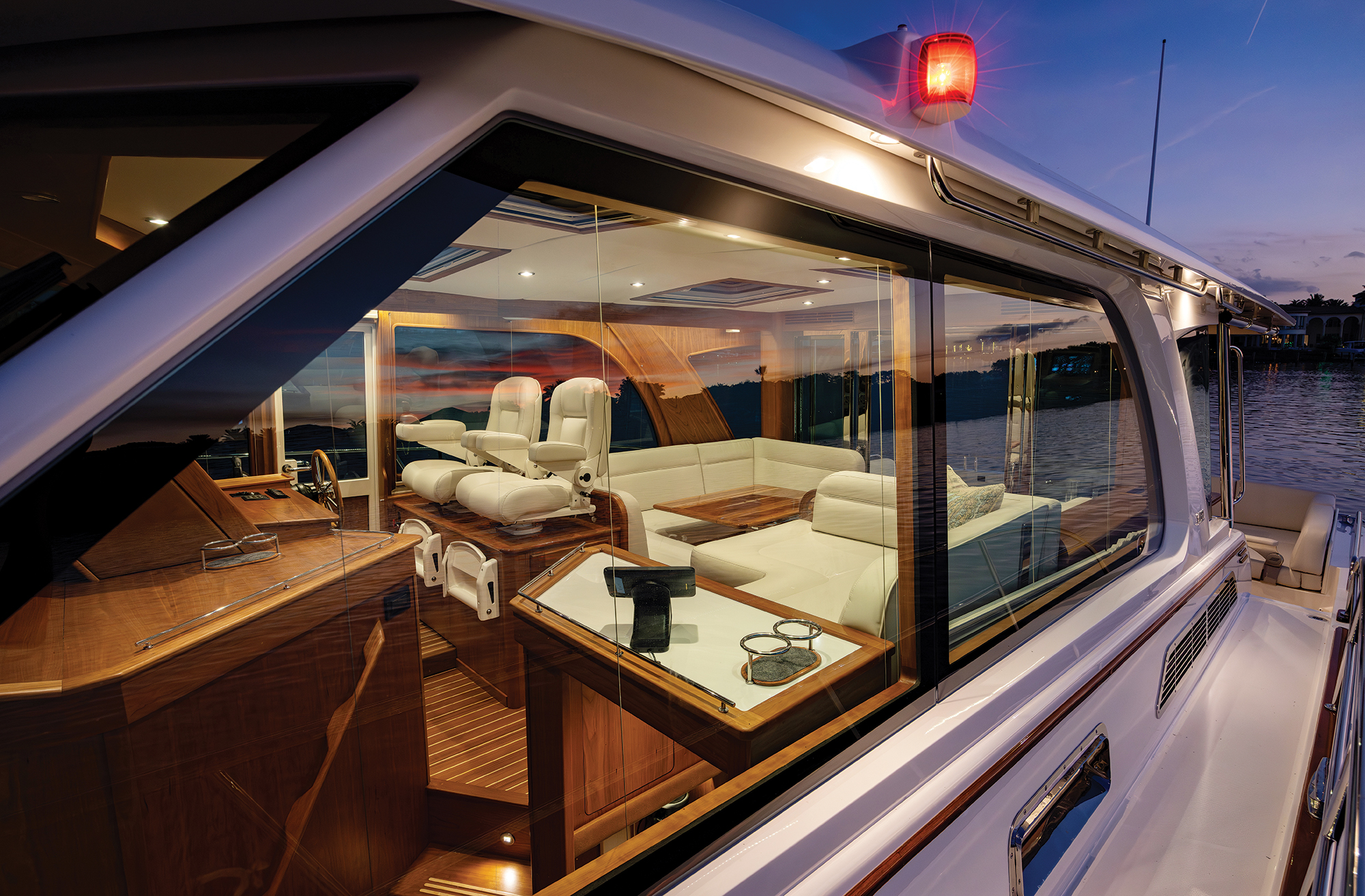An exterior dusk photograph looking through the large side windows into the illuminated, warm cherrywood salon and helm area of a Sabre motor yacht as the sun sets over the water.