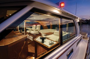 An exterior dusk photograph looking through the large side windows into the illuminated, warm cherrywood salon and helm area of a Sabre motor yacht as the sun sets over the water.