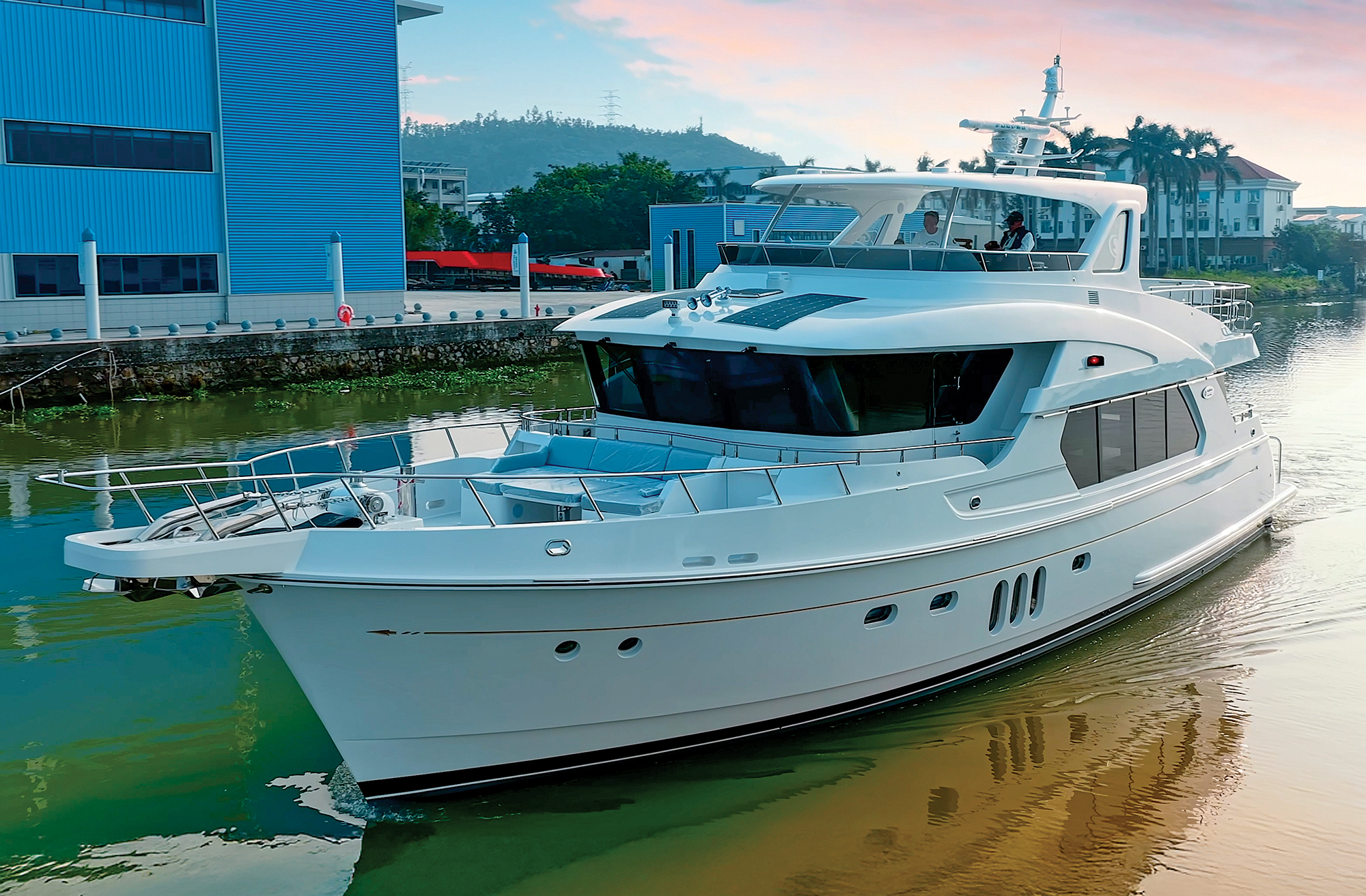 A forward three-quarter view of the white Selene 64 Ocean Explorer yacht navigating a canal alongside shipyard buildings under a soft twilight sky.