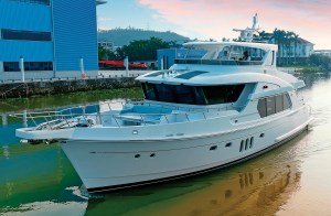 A forward three-quarter view of the white Selene 64 Ocean Explorer yacht navigating a canal alongside shipyard buildings under a soft twilight sky.