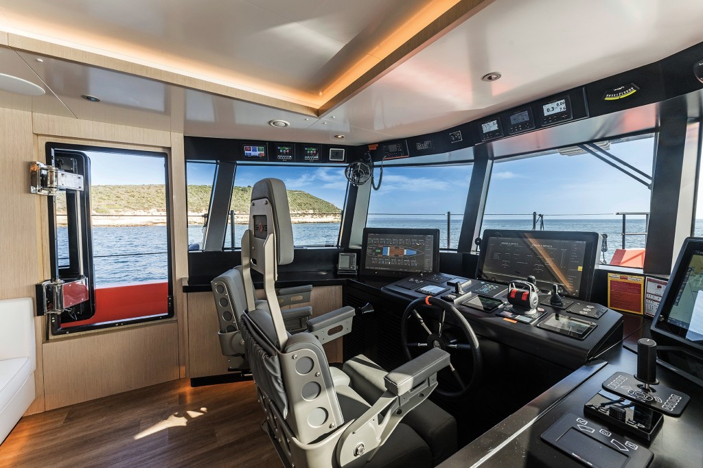 Modern helm station of the expedition yacht Vanguard featuring grey Shoxs suspension seating, a black steering wheel, multi-screen navigation displays, overhead digital instruments, and wide wraparound windows offering a clear view of open water and a coastal shoreline.
