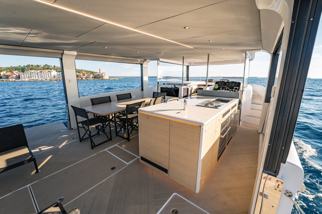 Open-air main deck salon of the Jeanneau Sea Loft 480 featuring a walkaround galley island, dining table with chairs, teak flooring, and large windows looking out at the ocean.