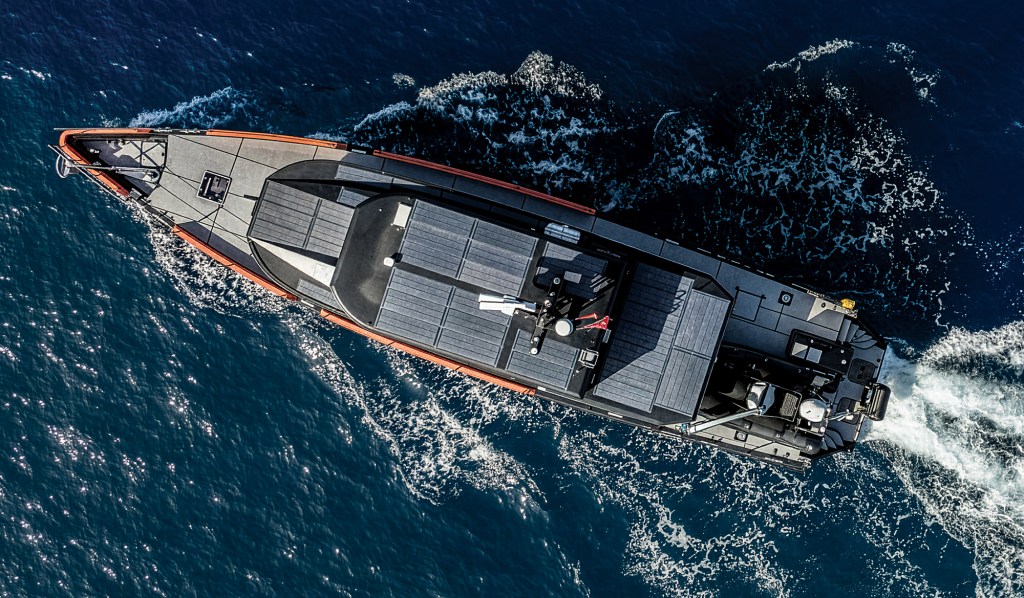 High-angle aerial view of the expedition yacht Vanguard cruising through deep blue water, showing its long orange hull, matte-black deck panels, rooftop solar panel array, and white wake trailing behind.