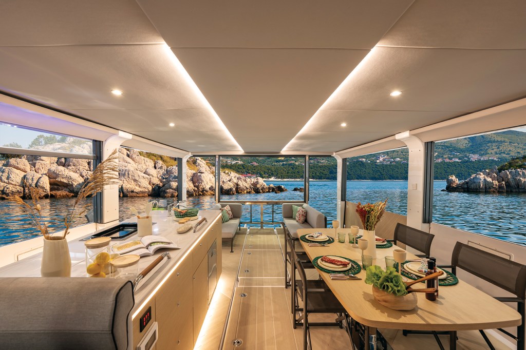 Interior view of the Jeanneau Sea Loft 480 salon at anchor, featuring a set dining table, kitchen island with warm strip lighting, aft lounge seating, and panoramic ocean views.