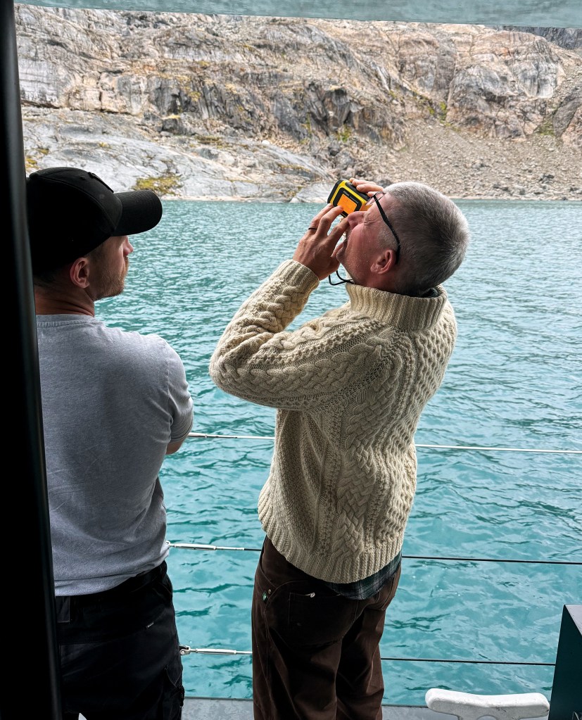 One man in a black cap watches while another in a thick, cream cable-knit sweater holds a small yellow electronic device up to his eyes like binoculars. The men are positioned on the right side of the frame, near the deck railings.