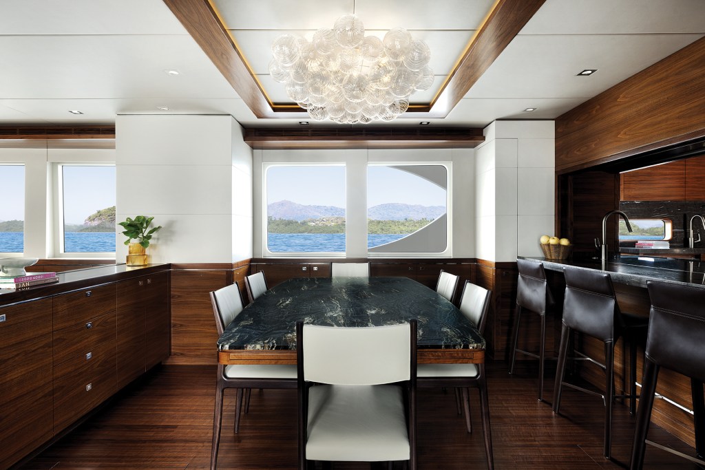 Interior dining area of the Nordhavn 100 yacht featuring a dark marble table, modern chandelier, bar seating, and large ocean-view windows.