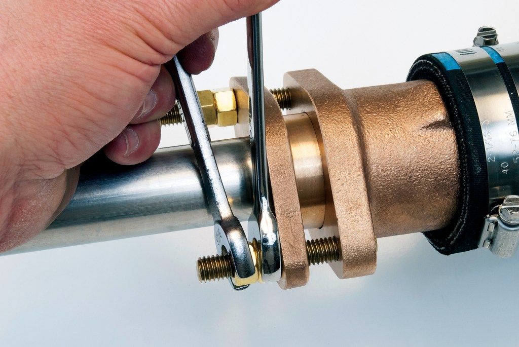 Hand using two open-end wrenches simultaneously on brass lock nuts of a boat propeller shaft stuffing box.