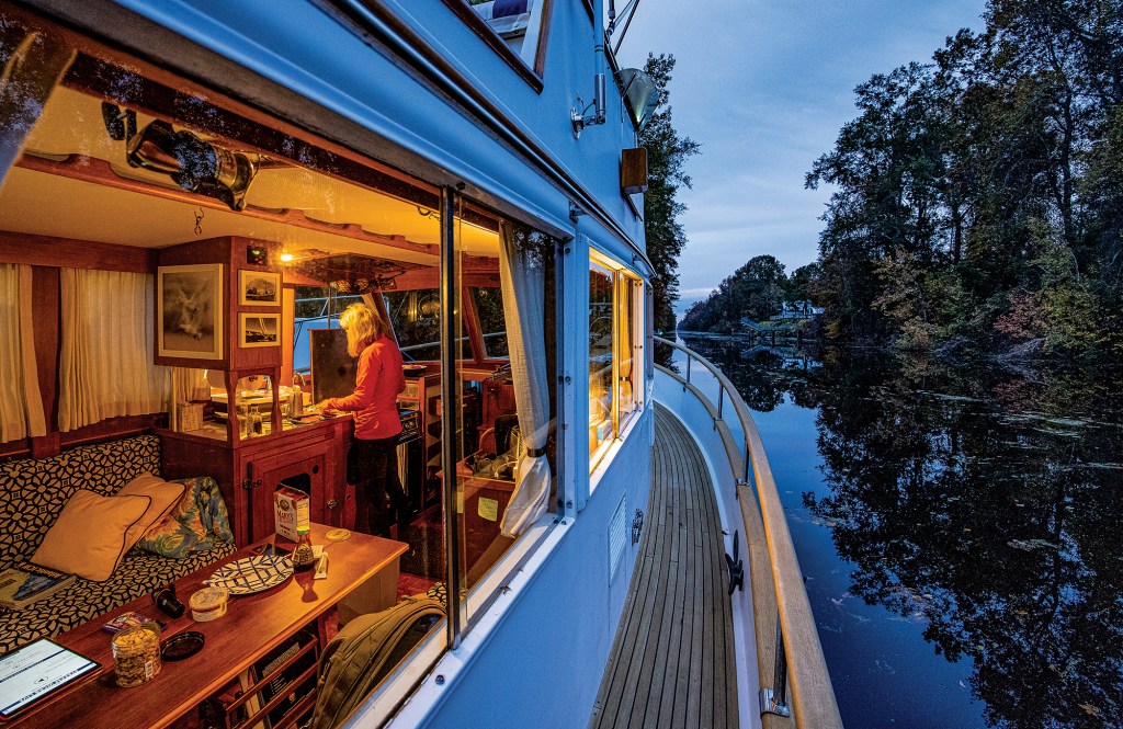Warmly lit interior cabin view of Tenley van der Wal preparing a meal aboard the trawler yacht Snow Goose at dusk.