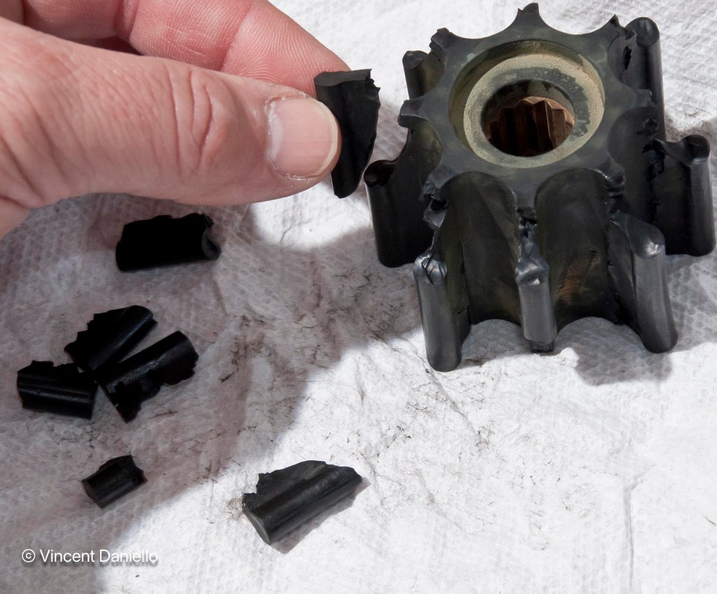 Close-up of a damaged black rubber marine water pump impeller with broken fins; several broken pieces are on the ground.