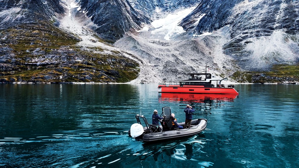 A wide landscape shot in a deep arctic fjord where crew members in a black Highfield RIB tender navigate calm, dark blue water in the foreground, with the bright orange custom expedition yacht Vanguard anchored nearby against towering, rugged, snow-dusted mountains.