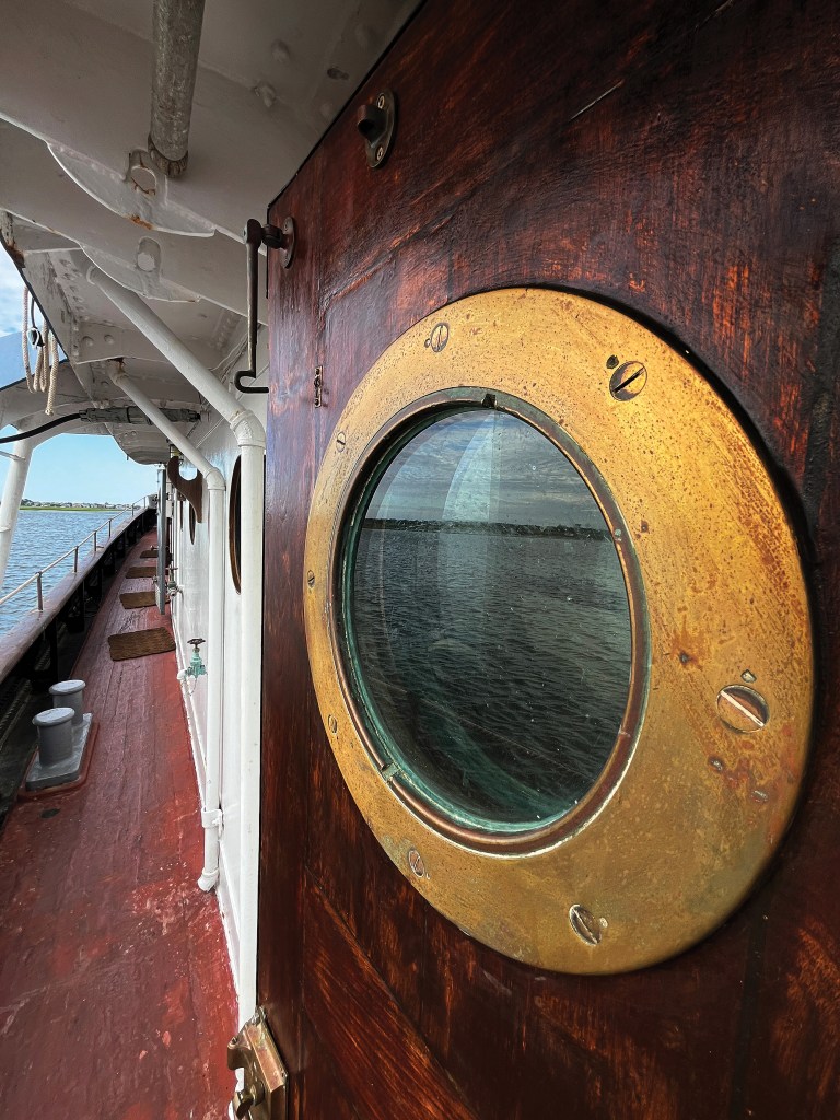 Brass porthole window on a wooden door along the side deck of the historic Robert Gray ship.