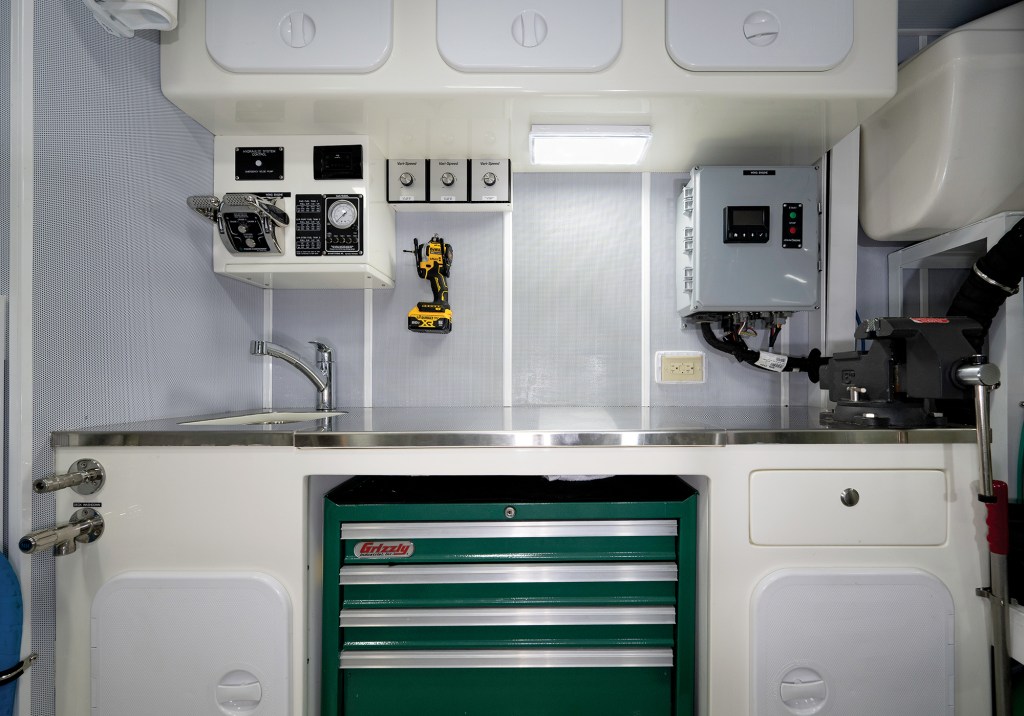 A fully equipped boat engine room workshop featuring a stainless steel workbench, bench vise, sink, wall-mounted DeWalt cordless drill, and built-in Grizzly tool chest.