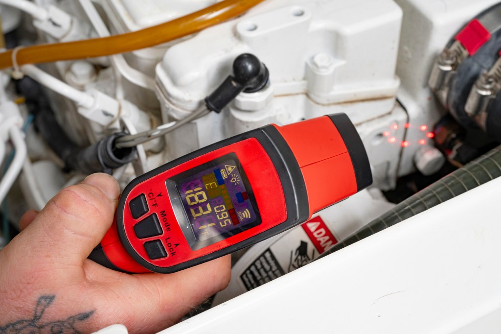 A hand holding a red infrared thermometer reading 183.1 degrees while aiming a red laser at a white boat engine component.