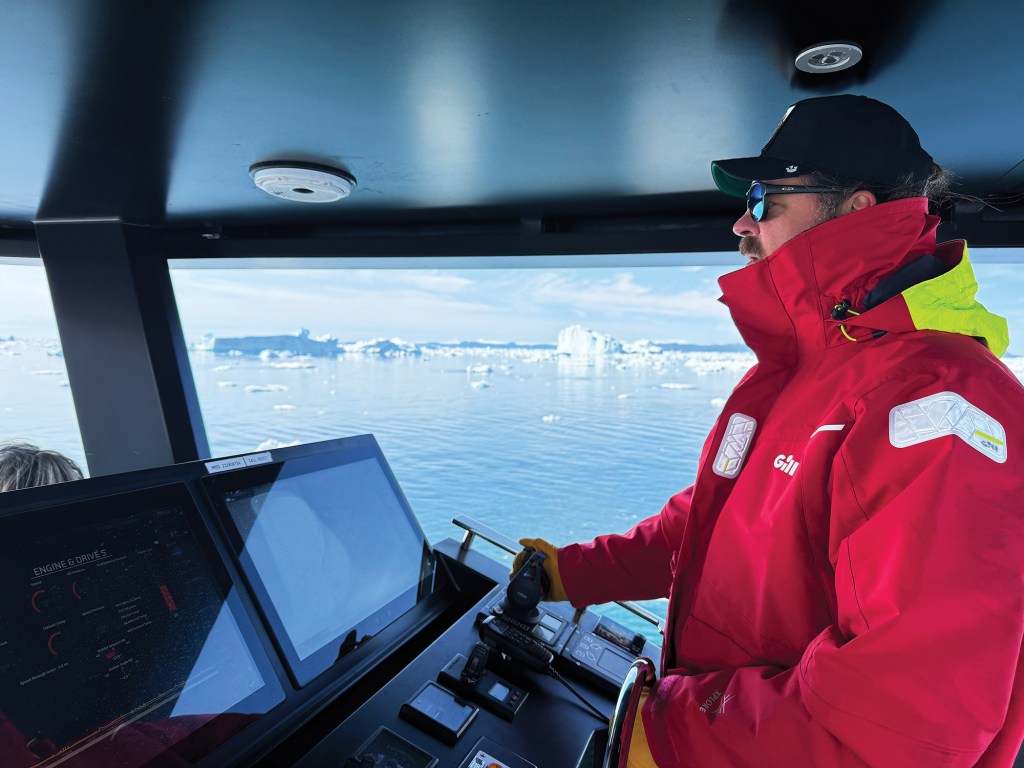 A man in profile wearing a red and high-vis yellow Gill foul-weather jacket, sunglasses, and a black cap stands at the helm of a yacht, navigating through an arctic waterscape dotted with distant icebergs seen through large cabin windows.