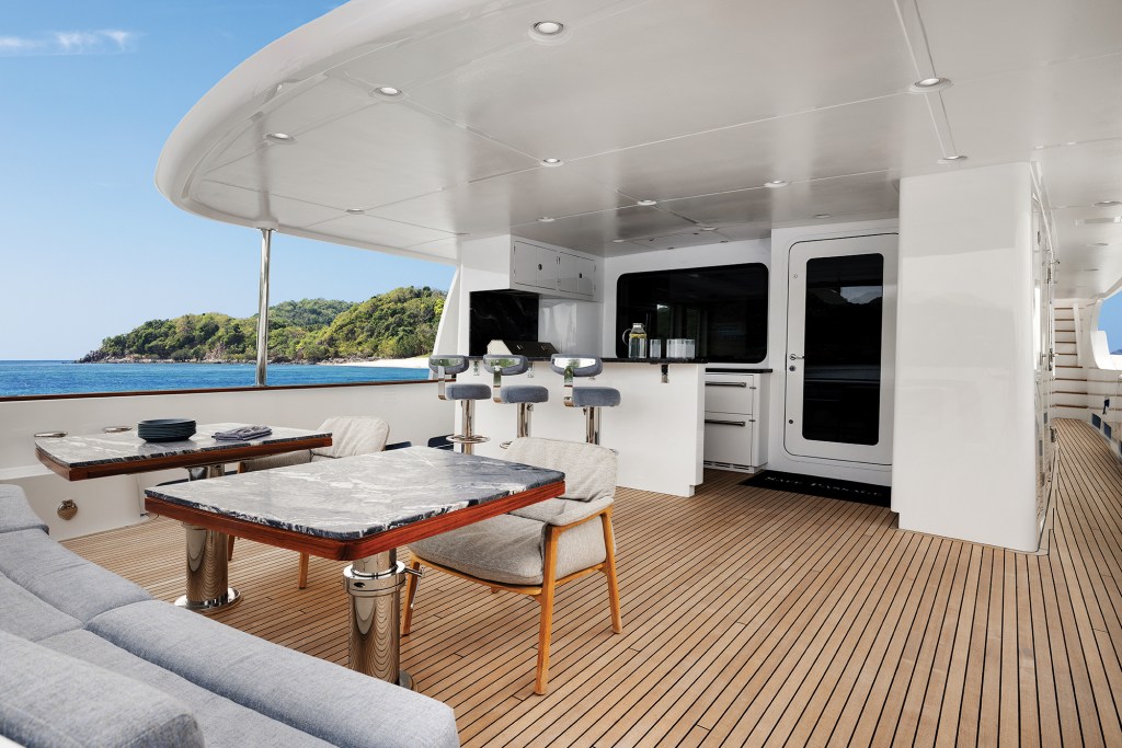 Covered aft deck on the Nordhavn 100 featuring teak flooring, outdoor dining tables, and a wet bar with seating overlooking the ocean.