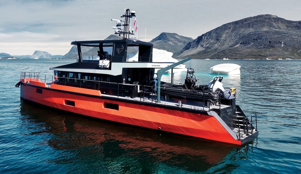 A rear-three-quarter view of the bright orange expedition yacht Vanguard sitting in calm water, showing its aft deck equipped with a crane and a black inflatable tender, with small icebergs floating nearby and dark, mountainous hills in the background.
