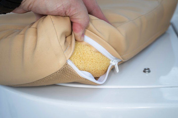 Hand unzipping a tan vinyl boat cushion showing reticulated foam padding and a breathable mesh bottom panel.