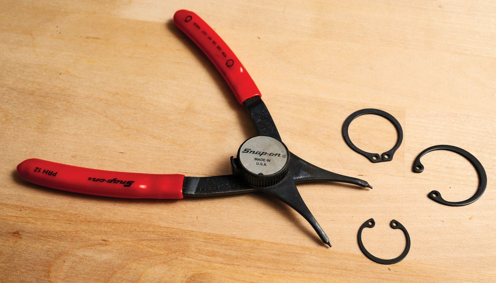 Red-handled Snap-on retaining ring pliers resting on a wooden surface next to three black snap rings.