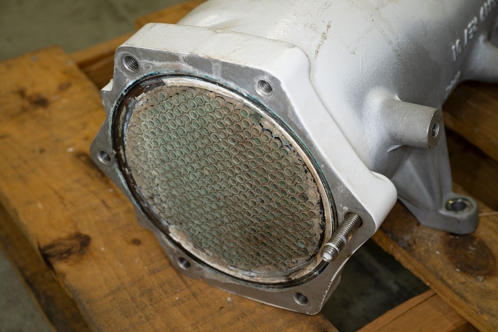 Close-up of a disassembled white marine heat exchanger showing the internal metal tube bundle grid resting on wood.