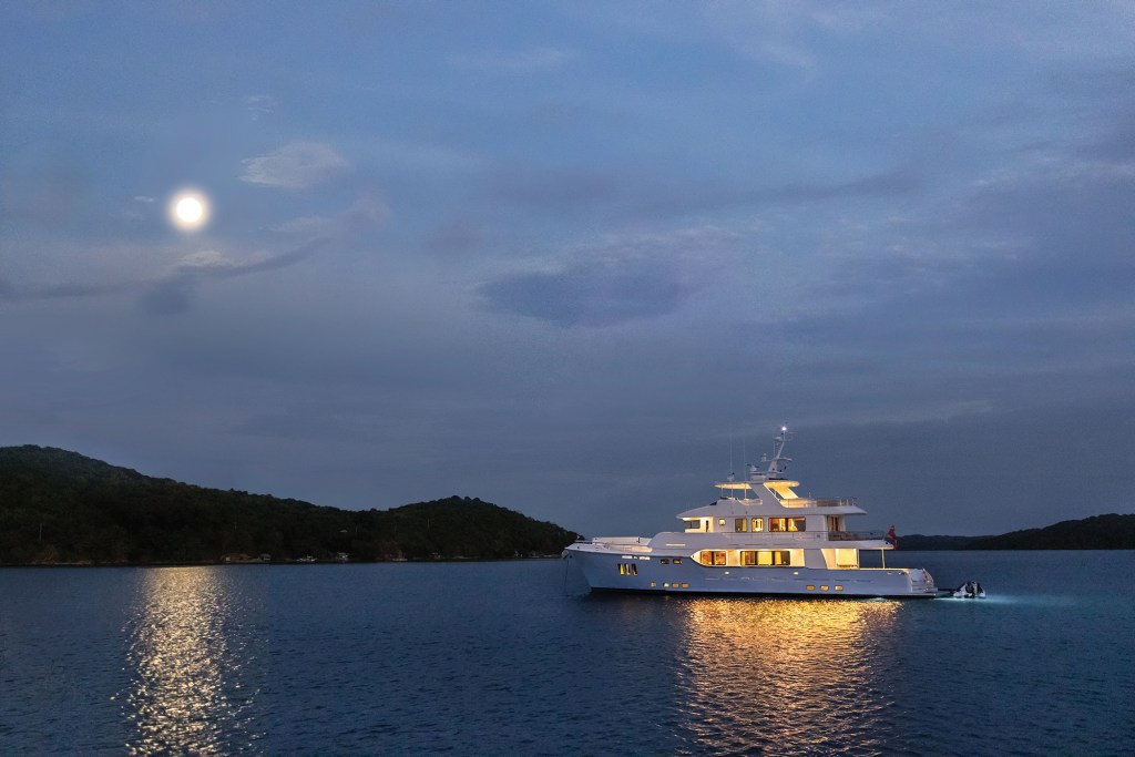 Nordhavn 100 luxury expedition yacht illuminated at anchor under a full moon in a quiet bay at dusk.