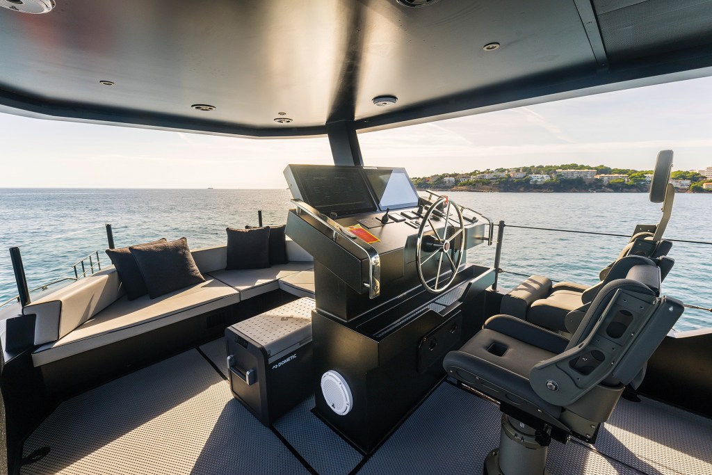 View of an open-air upper flybridge helm station on a yacht, featuring a central black console with a traditional steering wheel and navigation screens, grey Shoxs helm seats on the right, and a cushioned L-shaped bench seating area on the left overlooking sunny open water.