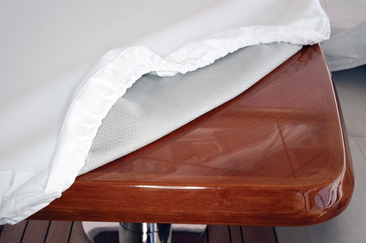Soft-lined protective white vinyl cover lifted to reveal a high-gloss varnished teak boat table corner.