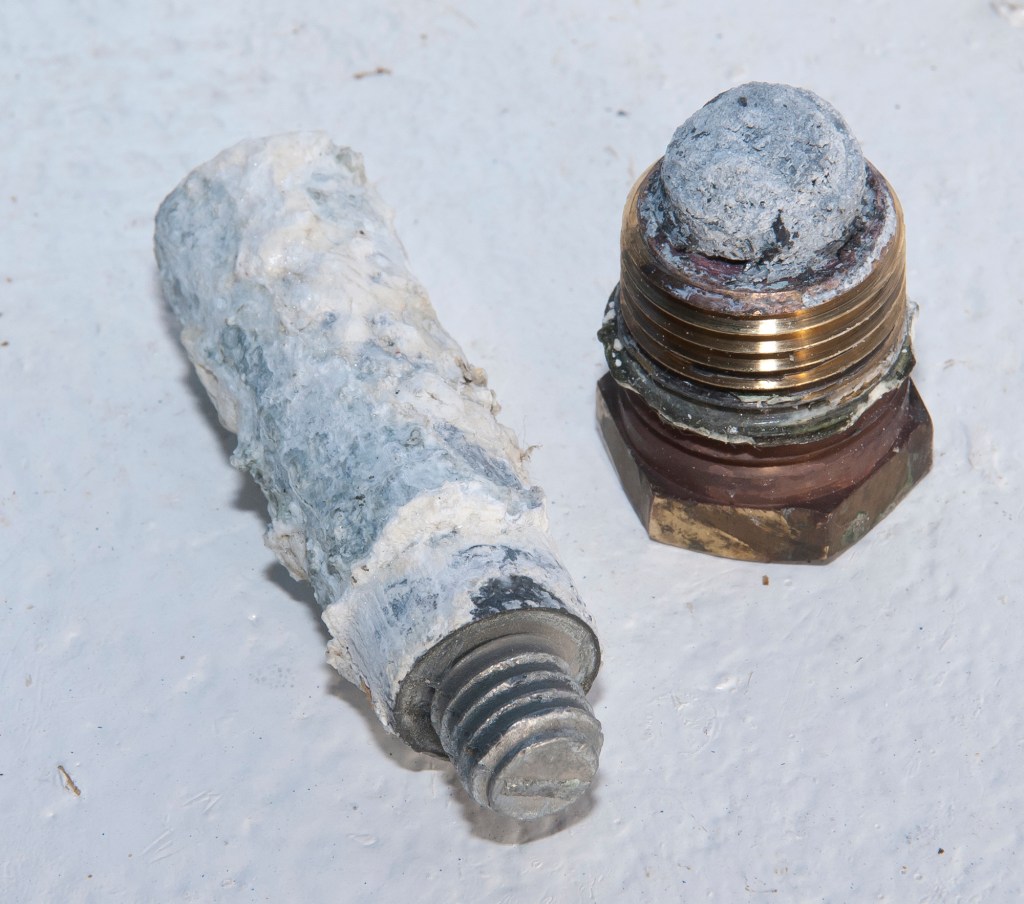 A new engine pencil zinc anode next to a heavily corroded, spent zinc plug on a light-colored surface.