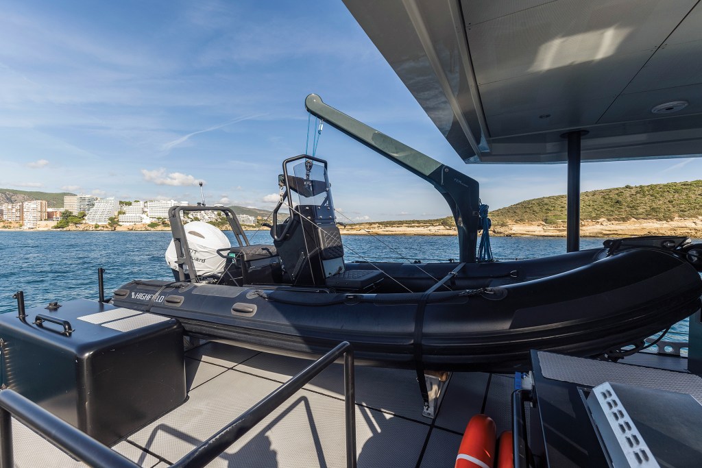 A matte-black Highfield RIB tender secured on the aft deck of a yacht under a dark davit crane arm, with grey Treadmaster deck flooring and a sunny coastal shoreline with buildings visible across blue water in the background.