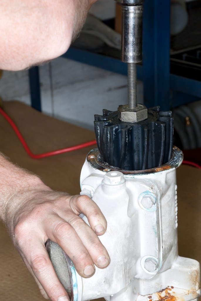 A mechanic using a specialized puller tool to remove a flexible black rubber impeller from a white pump housing.