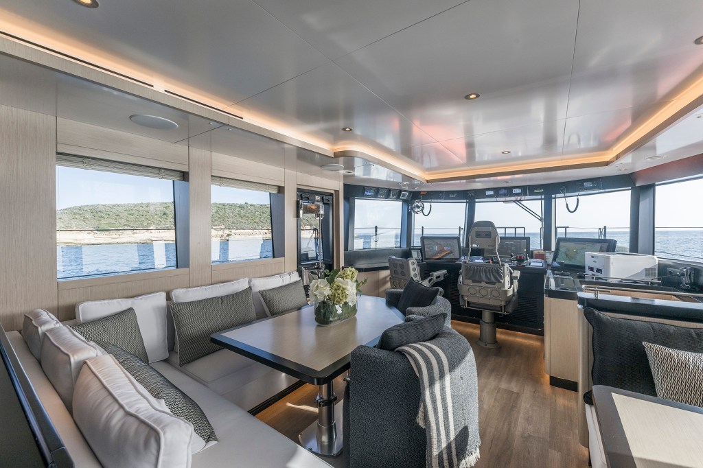 Main salon interior of a yacht looking toward the helm station, featuring soft light-wood wall paneling, a large dining table with a floral centerpiece, plush white and grey sofa seating, warm indirect ceiling lighting, and large windows looking out over water and coastline.