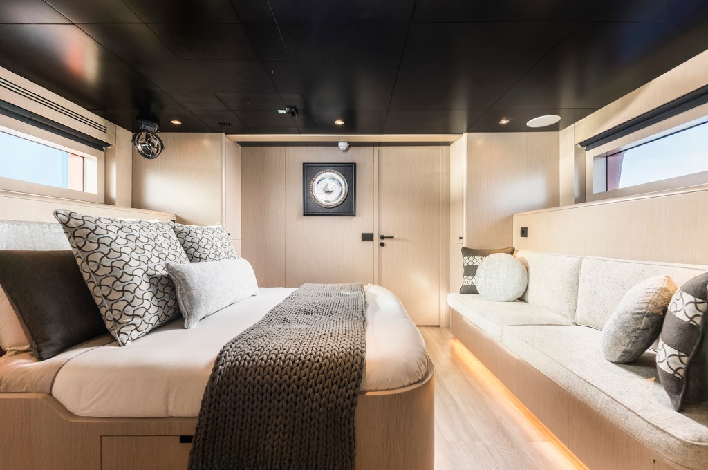 Modern master stateroom interior of a yacht featuring light-wood wall paneling, a dark ceiling, a king-size bed with geometric throw pillows and a grey knit blanket, and a built-in upholstered sofa running along the opposite wall under a long portlight window.