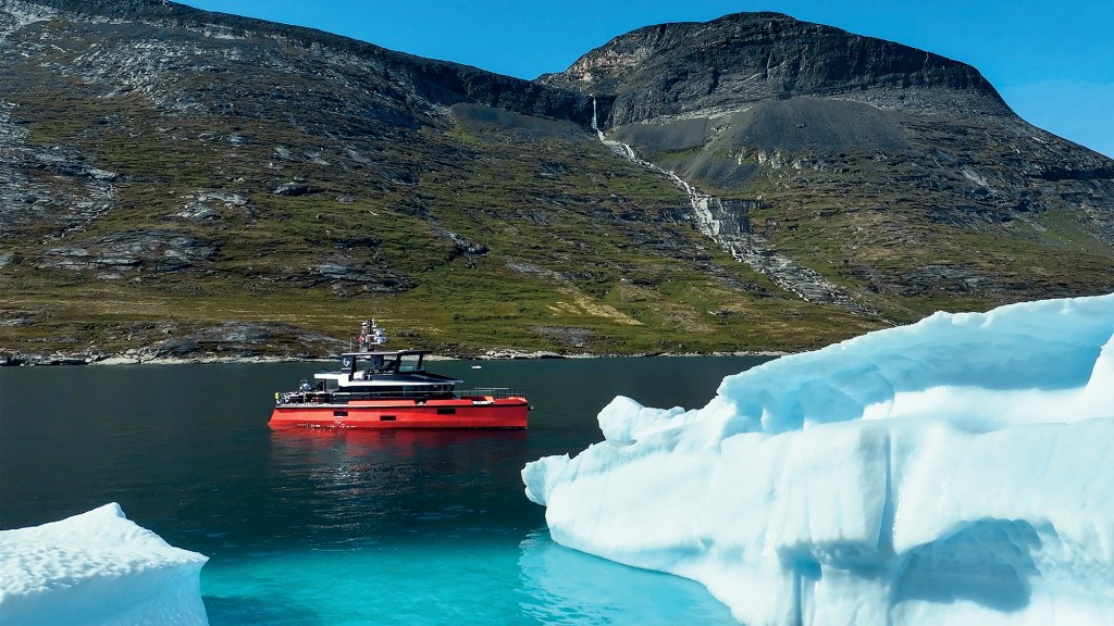A wide view of the orange expedition yacht Vanguard floating in deep, dark blue arctic waters framed by bright white icebergs in the foreground, with massive, rocky mountain slopes and a cascading waterfall in the background under a clear sky.