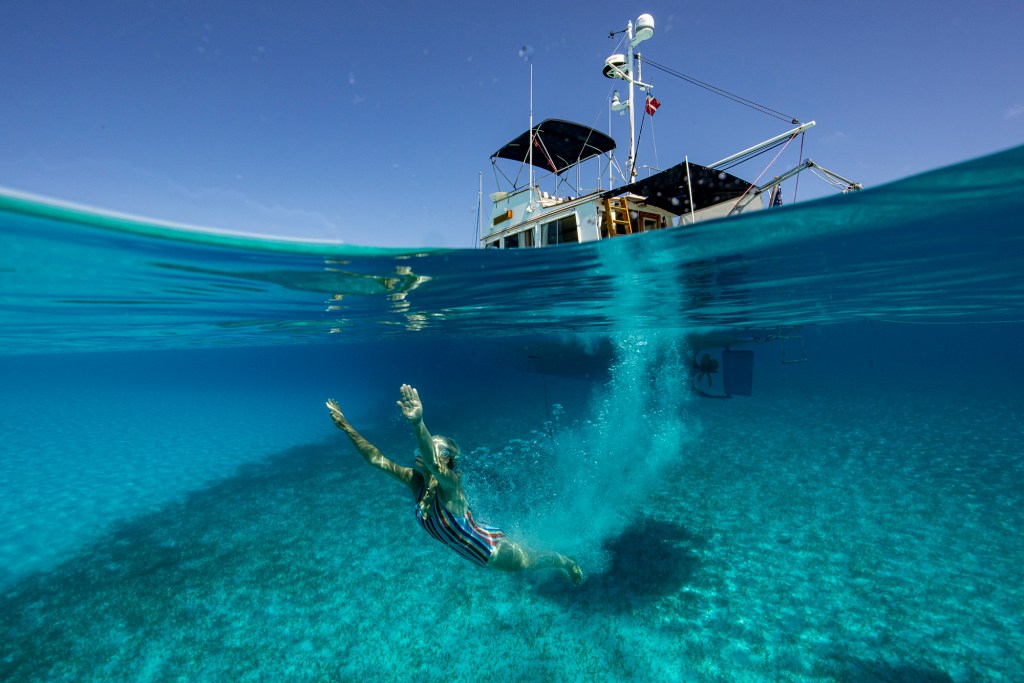 Split water-surface photograph of a swimmer diving underwater into clear tropical blue ocean next to the trawler yacht Snow Goose.