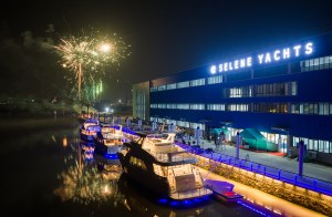 Nighttime view of the Selene Yachts shipyard and marina during a grand opening celebration, featuring a brilliant fireworks display bursting over the water, illuminate illuminated luxury yachts docked along blue LED-lit floating walkways alongside the main illuminated factory building.