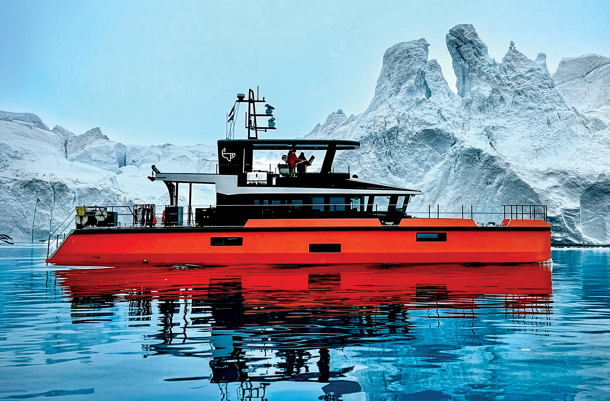 Side profile of the bright orange and black custom expedition yacht Vanguard floating in calm blue arctic waters, perfectly reflected on the surface against a backdrop of massive white and ice-blue glaciers under a pale blue sky.