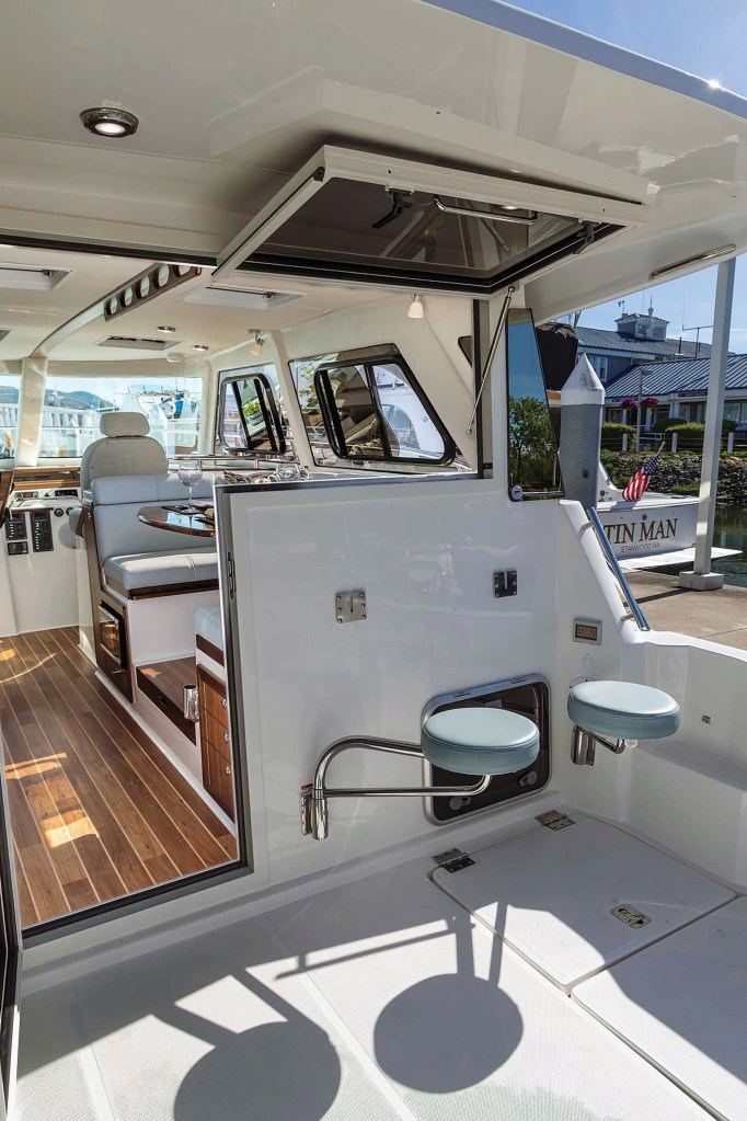 The aft cockpit of an Aspen C110 showing swing-out bar stools, a hinged bulkhead window open to the salon, and teak flooring indoors.