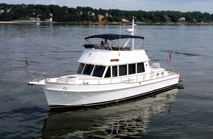 A white Grand Banks 47 Classic motor yacht named GiGi floating on calm waters in Hempstead Harbor, New York, with a dark blue flybridge bimini top and tree-lined shoreline in the background.